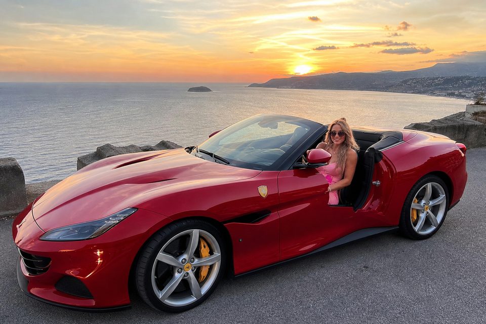Woman in sunglasses and a pink dress in a red convertible sports car at a seaside viewpoint at sunset