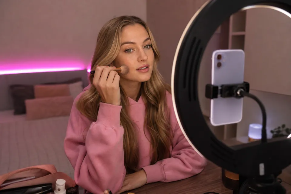Creator in a pink hoodie getting ready in front of a ring light with her phone mounted to film a promo clip